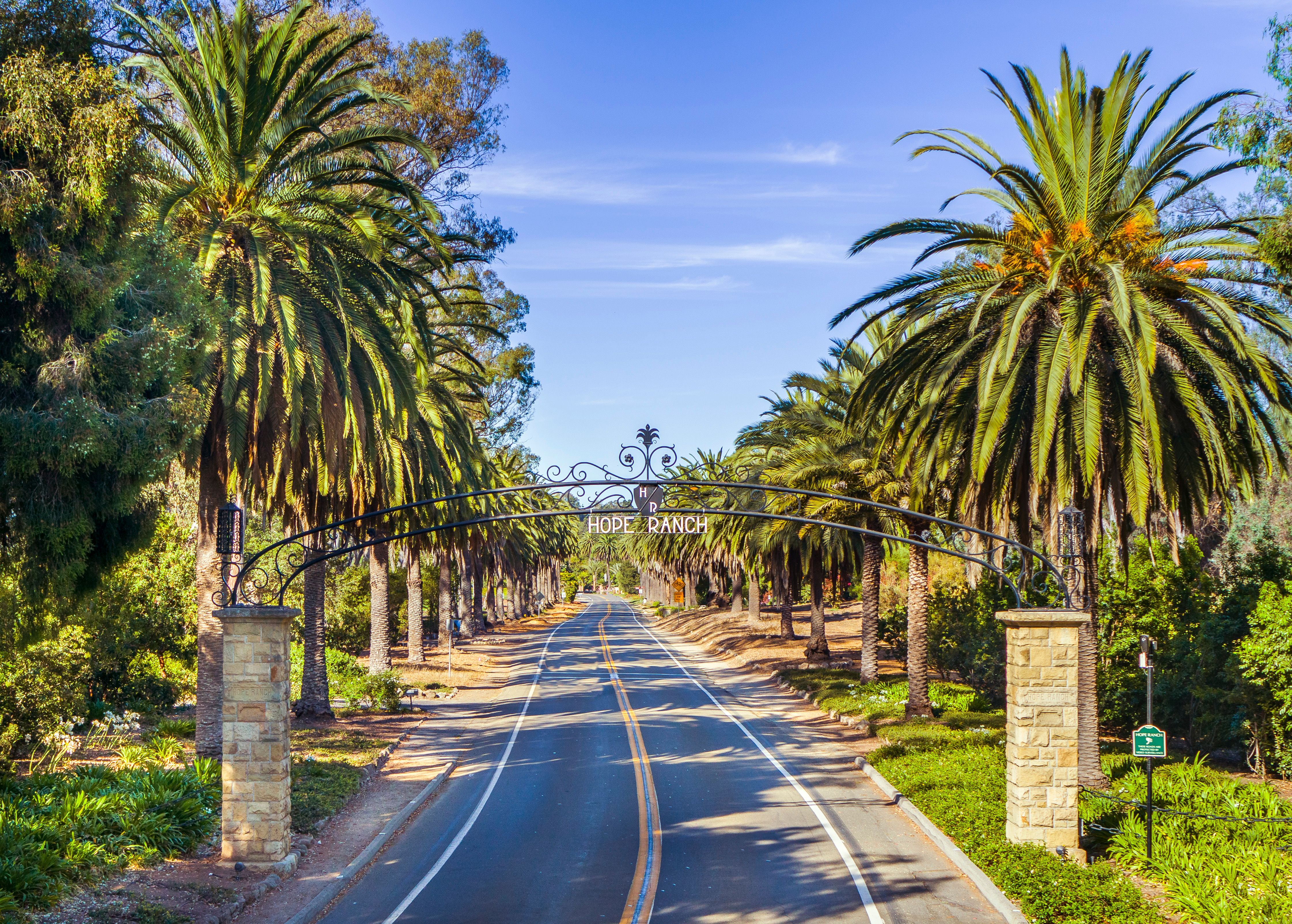 The paved road at the entrance to Hope Ranch in Santa Barbara, CA framed by palm trees, stone pillars, and a metal 